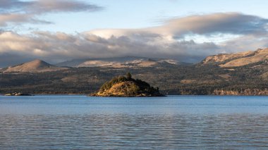 Ağaçlarla kaplı küçük bir göl, dağlarla kaplı bir gölde, Villa Pehuenia, Neuquen, Patagonya, Arjantin 'de bulutlu bir gökyüzü altında yer alır.