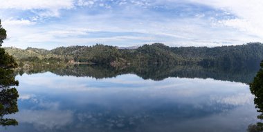 Sakin bir gölün panoramik manzarası ormanlı tepeleri ve Patagonya 'daki bulutlu gökyüzünü yansıtıyor.
