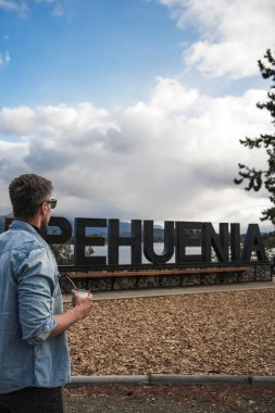 Turist adam elinde kupayla Villa Pehuenia 'ya doğru bakıyor. Arkasında göl ve dağlar olan hoş geldin tabelası var. 11 Nisan 2025 Neuquen, Patagonya, Arjantin