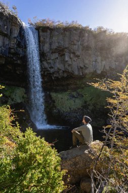 İnsan, sonbahar günışığı altındaki Cascada de la Vieja Usina şelalesine bakarak huzur içinde kayanın üzerinde oturur. Caviahue, Neuquen, Arjantin