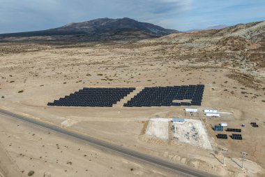 Kurak arazideki büyük bir güneş enerjisi santralinin havadan görünüşü. Parçalı bulutlu bir gökyüzünün altında dağlar var.