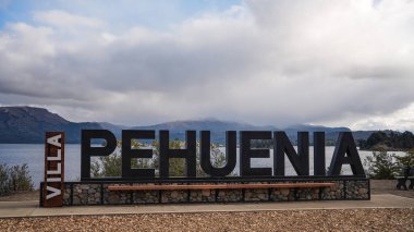 Büyük Villa Pehuenia karşılama tabelası Patagonya 'da bulutlu bir gökyüzünün altında dağları olan bir Alumine Gölü' nün yanında. 11 Nisan 2025. Villa Pehuenia, Arjantin