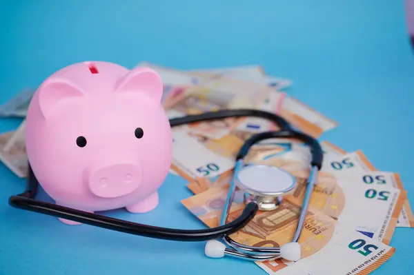 Çeşitli para birimleriyle çevrili bir kumbara ve sağlık harcamalarını gösteren bir steteskop.