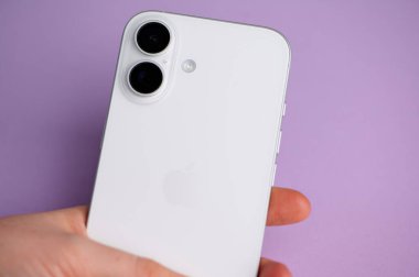 Mainz, Almanya - 09 Haziran 2025: kullanıcı yumuşak mor arkaplana karşı yerleştirilmiş çift kameralı parlak beyaz bir elma iPhone 16 tutuyor. Tasarım modern estetiği ve işlevselliği vurguluyor..