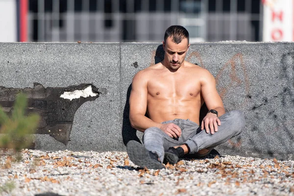 一个体格健壮的年轻人坐在古老的房顶上的画像 健美健美的肌肉健美模型在户外运动后的应用 免版税图库图片