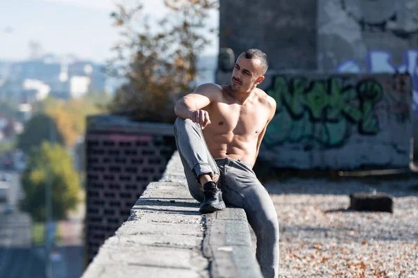 古い屋根の上に座っている彼のよく訓練された体を示す若い物理的にフィット男の肖像 運動後にポーズ筋肉運動体ビルダーフィットネスモデル屋外 ロイヤリティフリーのストック写真