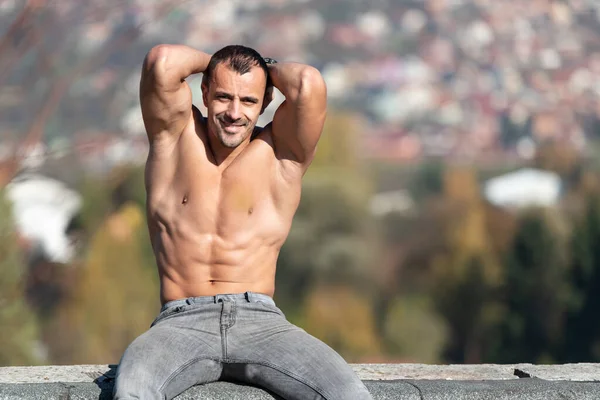 ハンサムな若い男は 古い屋根の上に強い屋外と曲げ筋肉に座って 筋肉運動ボディビルダーの男がポーズ ストック写真