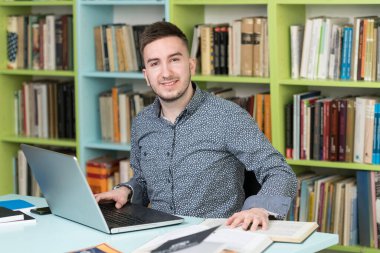 Kütüphanedeki Masada Oturan Yakışıklı Adam - Masadaki Dizüstü bilgisayar ve Düzenleyici - Arka Masadaki Bulanık Kitaplar