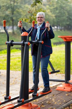 70 'li yaşlarda olgun bir kadın, huzurlu bir sonbahar parkı ortamında aktif kalmak için açık hava spor aletleri kullanır. Sıradan bir mavi kıyafet ve eşarpla, yaşlılara uygun olduğunu gösteriyor.