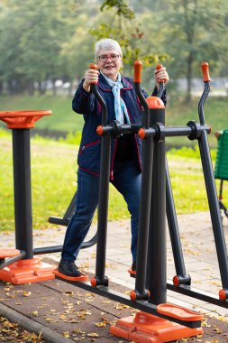 In a park filled with autumn colors, an elderly woman with glasses exercises on outdoor equipment. Her blue clothing and scarf complement the serene seasonal atmosphere clipart