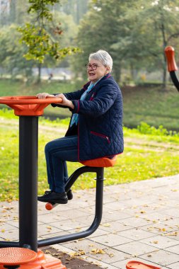 Bu görüntü, sonbahar kentsel parkında açık hava ekipmanları üzerinde egzersiz yapan gri saçlı bir kadını gösteriyor. Mavi elbisesi ve aksesuarları spor salonuna rahat ve sıradan yaklaşımını yansıtıyor.