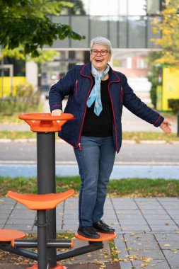 A senior woman in a blue jacket and scarf uses an exercise simulator in a public park. Her focus on fitness in an outdoor setting highlights healthy aging during the beautiful autumn season clipart