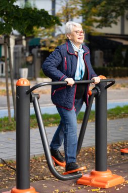 Gri saçlı ve günlük mavi elbiseli son sınıf öğrencisi bir kadın şehir parkında spor yapıyor. Forma odaklanmış durumda, yaşın sadece bir sayı olduğunu gösteriyor.