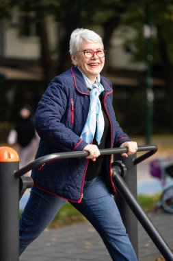 Sonbahar yeşillikleriyle çevrili bir şehir parkında, gözlüklü yaşlı bir kadın spor makineleriyle trenler kullanıyor. Mavi bir ceket ve eşarp giyiyor ve kendini sağlıklı bir yaşam tarzına adadığını gösteriyor.
