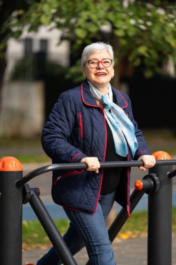 Bu görüntü, sonbahar kentsel parkında açık hava ekipmanları üzerinde egzersiz yapan gri saçlı bir kadını gösteriyor. Mavi elbisesi ve aksesuarları spor salonuna rahat ve sıradan yaklaşımını yansıtıyor.
