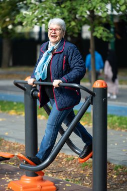 Beyaz saçlı, mavi-beyaz atkı takan bir kadın parktaki spor aletlerinin üzerinde, temiz sonbahar havasının tadını çıkarıyor. Aktif rutini sağlık ve sağlığa olan bağlılığını yansıtıyor.