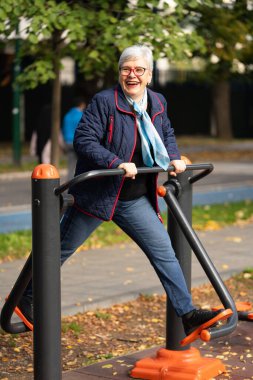 Gri saçlı ve günlük mavi elbiseli son sınıf öğrencisi bir kadın şehir parkında spor yapıyor. Forma odaklanmış durumda, yaşın sadece bir sayı olduğunu gösteriyor.