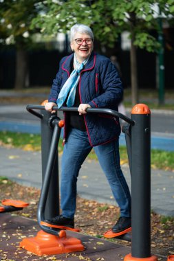 Sonbahar yeşillikleriyle çevrili bir şehir parkında, gözlüklü yaşlı bir kadın spor makineleriyle trenler kullanıyor. Mavi bir ceket ve eşarp giyiyor ve kendini sağlıklı bir yaşam tarzına adadığını gösteriyor.