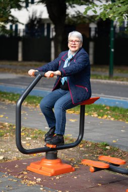 Şehir parkında spor makineleri üzerinde gri saçlı bir kadın sonbahar gününün tadını çıkarıyor. Günlük kıyafetlerle, mavi bir ceket, mavi pantolon ve atkı giyer, açık hava antrenmanına odaklanır.