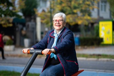 Sonbahar renkleriyle dolu bir parkta, gözlüklü yaşlı bir kadın açık hava ekipmanları üzerinde egzersiz yapıyor. Mavi elbisesi ve atkısı dingin mevsimlik atmosferi tamamlıyor.