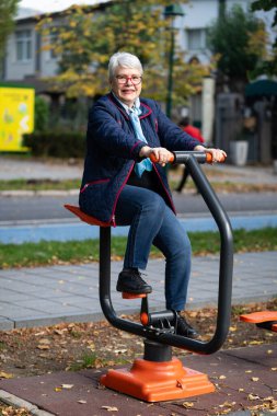 Bu görüntü, sonbahar kentsel parkında açık hava ekipmanları üzerinde egzersiz yapan gri saçlı bir kadını gösteriyor. Mavi elbisesi ve aksesuarları spor salonuna rahat ve sıradan yaklaşımını yansıtıyor.