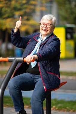 Şehir parkında spor makineleri üzerinde gri saçlı bir kadın sonbahar gününün tadını çıkarıyor. Günlük kıyafetlerle, mavi bir ceket, mavi pantolon ve atkı giyer, açık hava antrenmanına odaklanır.