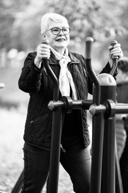 An older woman performs a workout on an exercise simulator in a park, dressed in blue clothing. With a casual outfit and glasses, she demonstrates active senior life in the autumn season clipart