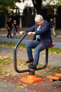 Sonbahar renkleriyle dolu bir parkta, gözlüklü yaşlı bir kadın açık hava ekipmanları üzerinde egzersiz yapıyor. Mavi elbisesi ve atkısı dingin mevsimlik atmosferi tamamlıyor.
