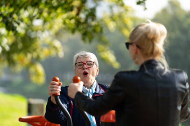 Anne ve kızı bir sonbahar parkında açık hava fitness makineleri üzerinde bir çalışma paylaşıyorlar. Mavili anne ve siyahlı kız şovdaki aile kondisyonu eğlenceli ve enerjik olabilir.