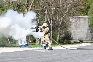 Sarı koruyucu giysili itfaiyeci dışarıdaki yanan metal bir yağ tenekesinde alevleri söndürüyor.
