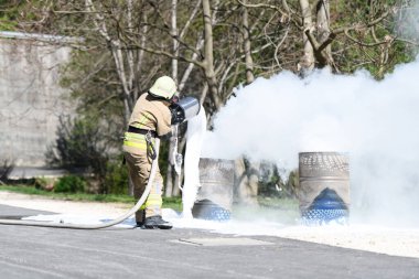 İtfaiyeci koruyucu kask takıyor. Yanan yağ tenekesini söndürmek için köpük püskürtüyor.