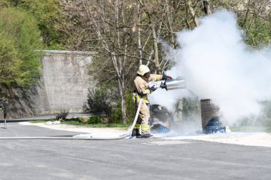 Sarı koruyucu giysili itfaiyeci dışarıdaki yanan metal bir yağ tenekesinde alevleri söndürüyor.