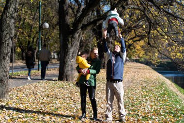 Ebeveynlerin ve küçük ikizlerin birlikte gülümsediği sonbahar parkı sahnesi.