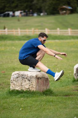 Parlak ve güzel bir günde çimenlerle ve ağaçlarla çevrili büyük bir taşın üzerinde çömelmiş tabancalarla pratik yapan bir adam.