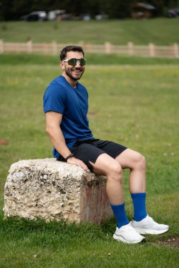 Güneş gözlüğü takan sporcu, arka planda çim ve ormanla manzaralı bir kayanın üzerinde otururken mola veriyor.