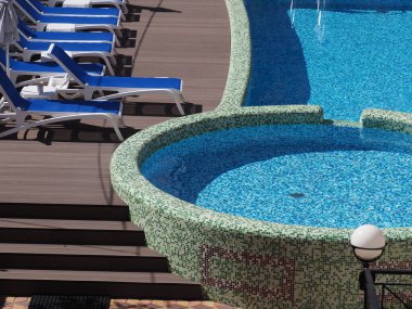 Mavi ve beyaz mozaikleri ve güneşli yatakları olan açık havada yuvarlak bir havuz.