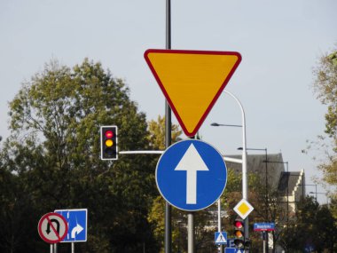 Arka planda kırmızı ışık ve ağaçların olduğu kavşakta yol işaretleri