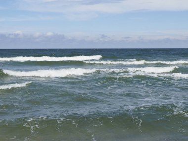 Denizde dalgalar mavi gökyüzünün altında, bulutlar kıyıdan görünüyor.