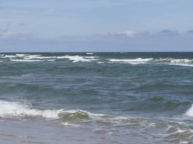 Mavi gökyüzünün altında kumlu bir sahilde dalgalı sakin bir deniz.