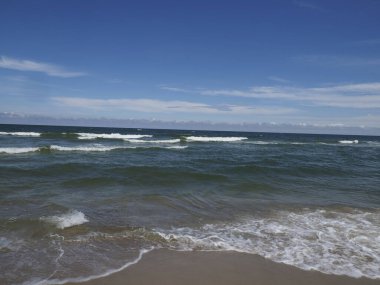 Sakin deniz manzarası, kumlu sahilde dalgalar mavi gökyüzü altında ve bir yaz gününde bulutlar.
