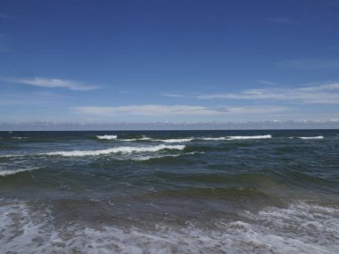 Mavi gökyüzünün altında dalgalar ve ufukta beyaz bulutlar olan sakin bir deniz.