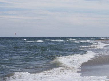 Mavi gökyüzünün altında dalgalar ve uçurtmalarla kumsalda