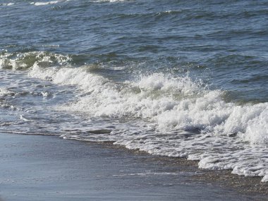 Dalgalar güneşli bir gökyüzünün altında kumlu bir plaja çarpıyor