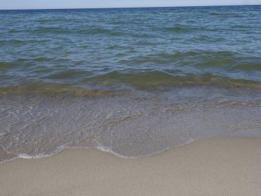 Sakin deniz, berrak gökyüzünün altında kumlu sahil, dalgalar kıyıya vuruyor.