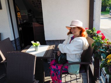 Beyaz ceketli ve pembe şapkalı bir kadın açık bir kafe masasında çiçeklerle oturuyor.