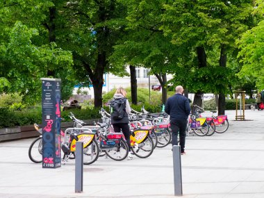 İnsanlar yeşil ağaçların altına park etmiş renkli bisikletleri olan şehir bisiklet kiralama istasyonunu kullanıyorlar.