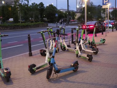 Şehir ışıklarıyla şehir kaldırımına park etmiş renkli elektrikli scooterlar. Arka planda binalar var.