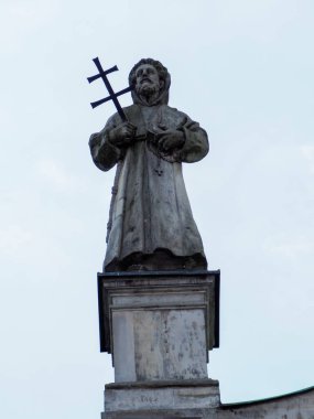 Karanlık bronz dini heykel, anıt düzeninde bulutlu gökyüzüne karşı haç tutuyor.