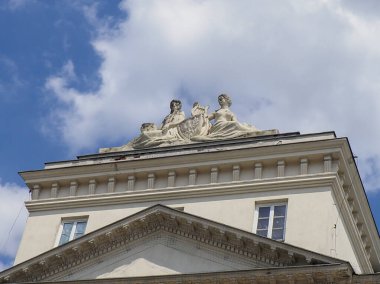 Neoklasik yapı köşesinde heykel figürleri ve dramatik bulutlu mavi gökyüzüne karşı üçgen pedikürasyon ayrıntıları yer alıyor