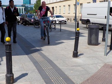 Modern sokak mobilyaları ve dokunsal kaldırım sisteminin yanında yürüyen ve bisiklete binen insanların bulunduğu kentsel yaya bölgesi